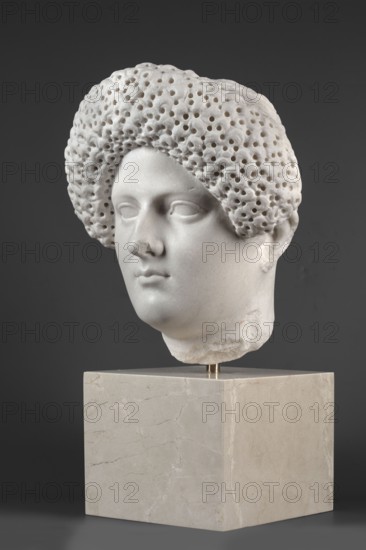 Portrait of a Roman woman, head with a broad face and an elaborate hairstyle: hair twisted into small curls marked with a gimlet, the rest gathered at the back in a small bun; round, full face with a triangular forehead. Female portrait, Roman sculpture, masterpiece; Roman Empire 27 BC–476, ca. 82–92, Rome. Sculpture. Height 30.5 cm; width 27 cm; depth 25 cm; weight 36.5 kg., A white marble sculpted head with textured, perforated hair displayed on a pale beige marble block against a dark gray background, showing smooth facial features including closed eyes, a straight nose, and full lips, with subtle shadows and highlights on the pale stone surface and a few small rough or chipped areas on the neck and base.