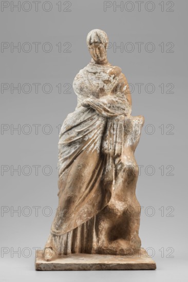 Female figurine. The figurine depicts a standing woman wrapped in a cloak, leaning her left elbow against a rock. Traces of paint have been preserved: red on the sandal worn by the figure's left foot and on the lips, white on the cloak, and blue on the chiton, a fragment of which peeks out from beneath the outer garment. The modeling of the figure is very precise and accurate, particularly the face and the slightly tilted right head, covered by the cloak. The position of the body leaning against the rock—slightly bent—with crossed legs, clearly outlined under the cloak the left leg is moved forward and the aforementioned tilt of the head in the opposite direction to the rest of the body give the figurine a melancholic mood. 4th century BC, Tanagra Greece. Figurine; height 35.5 cm, width 16.5 cm, depth 13.8 cm, weight 1.45 kg., A standing sculpture of a draped human figure with a rounded head and short hair, wearing flowing garments that form layered folds and a wrapped mantle, leaning ...