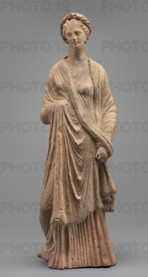 Female Figurine, Standing woman dressed in a long robe, slightly pleated and belted high under the breasts. The cloak, opened slightly at the front, falls below the knees, covering the arms except for the left hand, which holds the cloak. The body weight rests on the left leg, the right is pulled back. The head is modeled very carefully, the hair arranged in waves, with a gilded wreath atop it. Earrings in the ears. 2nd century BC, Myrina. Terracotta figurine. Height 31.5 cm, width 10.9 cm, depth 10 cm, weight 0.83 kg., A weathered standing female figure carved from a beige-brown material, shown full-length against a neutral gray background; she wears a draped garment with flowing folds and pleats, a shawl wrapped around her shoulders and across her torso, and a braided headband; her facial features are worn but show a calm expression, and one arm is bent bringing the drapery across her waist while the other hangs at her side holding fabric; the surface shows rough texture and slight d...