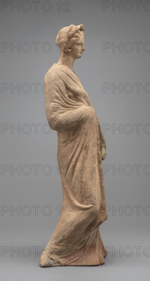 Female figurine, standing woman dressed in a long robe, slightly pleated and belted high under the breasts. The cloak, opened slightly at the front, falls below the knees, covering the arms except for the left hand, which holds the cloak. The body weight rests on the left leg, the right is pulled back. Head modeled carefully, hair arranged in waves, with a gilded wreath atop it. Earrings in the ears. Terracotta figurine, 2nd century BC, from Myrina. Height 31.5 cm, width 10.9 cm, depth 10 cm, weight 0.83 kg., A standing terracotta figure in beige and light brown tones draped in a flowing garment with visible folds, shown in profile facing right, with a head wrapped in a cloth or band and detailed facial features, arms positioned with one bent and the other resting forward beneath the drapery, the surface showing textured weathering and small darker specks, set against a smooth gray background.