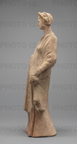 Female figurine: standing woman dressed in a long robe, slightly pleated and belted high under the breasts. The cloak, opened slightly at the front, falls below the knees, covering the arms except for the left hand, which holds the cloak. The body weight rests on the left leg, the right is pulled back. The head is modeled very carefully, the hair arranged in waves, with a gilded wreath atop it. Earrings in the ears. Terracotta figurine, 2nd century BC, Myrina. Height 31.5 cm; width 10.9 cm; depth 10 cm; weight 0.83 kg., A standing terracotta figure viewed in profile, rendered in beige and light brown tones with traces of darker brown and faint gold on a headband; the figure wears a long draped garment with visible folds and flowing lines, hands clasped at the front, a rounded base, and a smooth neutral gray background.