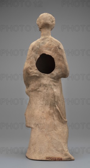 Female Figurine. Standing woman dressed in a long robe, slightly pleated and belted high under the breasts. The cloak, opened slightly at the front, falls below the knees, covering the arms except for the left hand, which holds the cloak. The body weight rests on the left leg, the right is pulled back. The head is modeled very carefully; the hair arranged in waves with a gilded wreath atop it. Earrings in the ears. 2nd century BC, Myrina. Terracotta figurine. Height 31.5 cm, width 10.9 cm, depth 10 cm, weight 0.83 kg., A standing figure sculpture viewed from the back, made of a rough-textured material in tan and beige tones with darker brown and gray mottling, showing a rounded head and draped clothing with visible folds and creases, a large circular hollow in the upper back, and a flat base in similar tan color; the background is a smooth medium gray.