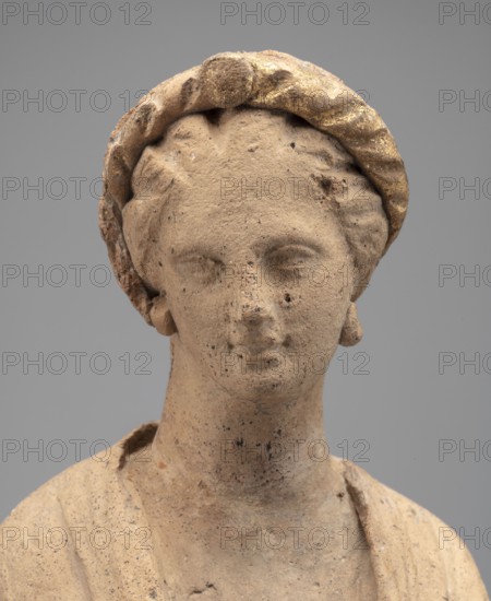 Female Figurine. Standing woman dressed in a long robe, slightly pleated and belted high under the breasts. The cloak, opened slightly at the front, falls below the knees, covering the arms except for the left hand, which holds the cloak. The body weight rests on the left leg, the right is pulled back. The head is modeled very carefully, the hair arranged in waves, and a gilded wreath atop it. Earrings in the ears. Terracotta figurine, 2nd century BC, Myrina. Height 31.5 cm; width 10.9 cm; depth 10 cm; weight 0.83 kg., A close-up view of a weathered terracotta head and upper torso of a figure with a rounded face and serene expression, wearing a textured headband with traces of gold and a pair of small, rounded earrings; the surface shows pitting and small dark speckles, with the terracotta displaying warm beige and light brown tones contrasted by the muted gold on the headband, set against a smooth neutral gray background.