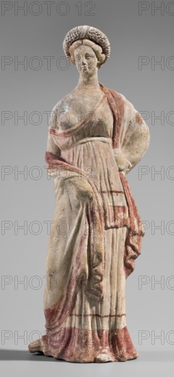 Figurine of a maenad, ca. 3rd–2nd century BC. A young woman standing in a wreath tied with a ribbon is dressed in a chiton slipped from her right shoulder exposing her right breast and upper arm, and a cloak that envelops her hands. The left hand rests on her hip; the right leg is pushed back. The robe is light pink with a wide border at the bottom and a narrow beetroot-colored stripe above it; a similar wide border runs around the cloak. The back of the figurine is unfinished and it has no base. Provenance: Myrina. Terracotta figurine; height 26.5 cm, width 9.2 cm, depth 7.4 cm, weight 0.39 kg., A standing statue of a robed female figure with a braided or coiled hairstyle, rendered in a weathered pale stone with visible areas of faded red paint on the drapery and trim; the figure wears a long, pleated dress with a draped shawl or cloak over one shoulder, one arm bent behind the back and the other hand holding a fold of fabric; the face is worn with a neutral expression, the surface sh...