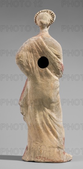 Figurine of a maenad. A young woman standing in a wreath tied with a ribbon is dressed in a chiton slipped from her right shoulder exposing her right breast and upper arm, and a cloak that envelops her hands. The left hand rests on her hip; the right leg is pushed back. The robe is light pink and decorated at the bottom with a wide border and a narrow beetroot-colored stripe above it. A similar border is found at the bottom of the cloak, and a wide border in the same color runs around the cloak. The back of the figurine is unfinished. Without base. 3rd–2nd century BC previously dated 2nd century BC. Myrina. Figurine. Height 26.5 cm; width 9.2 cm; depth 7.4 cm; weight 0.39 kg., A worn statue viewed from the back showing a standing human figure draped in flowing garments with visible folds and a rounded halo-like element around the head; the surface shows a patina of beige and tan with areas of faded reddish pigment and small darker spots, and there is a dark circular hole in the upper b...
