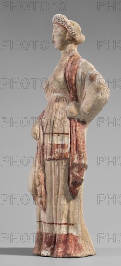 Figurine of a maenad. A young woman standing in a wreath tied with a ribbon is dressed in a chiton slipped from her right shoulder so as to expose her right breast and upper arm, and a cloak that envelops her hands. The left hand rests on her hip; the right leg is pushed back. The robe is light pink and decorated at the bottom with a wide border and a narrow beetroot-colored stripe above it. A similar border is found at the bottom, and a wide border in the same color runs around the cloak. The back of the figurine is unfinished. Without base. 3rd–2nd century BC previously dated 2nd BC, Myrina findspot, figurine, height 26.5 cm, width 9.2 cm, depth 7.4 cm, weight 0.39 kg., A weathered standing figure of a woman seen in profile with a coiffed hair arrangement, wearing a long draped dress and a shawl; the surface shows worn pigment in shades of pale beige, off-white, reddish-brown, and muted pink, with traces of darker brown in folds and creases; the figure's left arm rests on the hip whi...