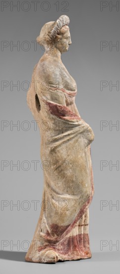 Figurine of a maenad. A young woman standing in a wreath tied with a ribbon is dressed in a chiton slipped from her right shoulder exposing her right breast and upper arm, and a cloak that envelops her hands. The left hand rests on her hip; the right leg is pushed back. The robe is light pink decorated at the bottom with a wide border and a narrow beetroot-colored stripe above it. A similar border is found at the bottom, and a wide border in the same color runs around the cloak. The back of the figurine is unfinished. Without base. 3rd–2nd century BC. Myrina. Terracotta figurine. Height 26.5 cm, width 9.2 cm, depth 7.4 cm, weight 0.39 kg., A standing statue of a woman shown in profile facing right, rendered in a weathered pale beige material with traces of muted red pigment on the drapery and hair, wearing a long flowing garment with visible folds gathered at the waist; the figure has coiled hair or a braided headpiece, a defined nose and chin, and a rounded shoulder with an opening at...