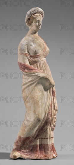 Figurine of a maenad. A young woman standing in a wreath tied with a ribbon is dressed in a chiton slipped from her right shoulder to expose her right breast and upper arm, and a cloak that envelops her hands. The left hand rests on her hip; the right leg is pushed back. The robe is light pink, decorated at the bottom with a wide border and a narrow beetroot-colored stripe above it. A similar border is found at the bottom, and a wide border in the same color runs around the cloak. The back of the figurine is unfinished. Without base. 3rd–2nd century BC previously dated 2nd BC. Myrina; terracotta figurine; height 26.5 cm, width 9.2 cm, depth 7.4 cm; weight 0.39 kg., A standing statue of a woman with a rounded, textured headdress and an upturned face, rendered in weathered pale stone with traces of reddish-pink pigment on the drapery and across an arm, a pale beige body with surface discoloration and small dark speckles, and a neutral gray background; she wears a flowing garment gathered...