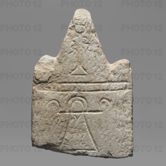 Votive stele with the sign Tanit, unknown Carthaginian workshop. Upper part of a stele with acroteria. The triangular pediment of the stele is separated from its shaft by a schematic frieze composed of four triglyphs and four metopes. In the triangular field is an engraved representation of a banner with ribbons, placed between the acroteria. In the centre of the stele is a representation of a sign interpreted as the sign of the goddess Tanit; on the pediment a banner. All decoration made with engraved lines. 3rd–2nd century BC. Carthage place of finding and origin. Stele; height 15.5 cm, width 10.8 cm, depth 3.9 cm, weight 0.99 kg., A roughly triangular stone slab with a textured, weathered surface in shades of gray and beige, carved with simple geometric incisions including a large triangle near the bottom, a horizontal band above it, and a few arch-like curved lines; the top narrows to a pointed peak with irregular edges and a small carved circular shape near the summit, and the sto...