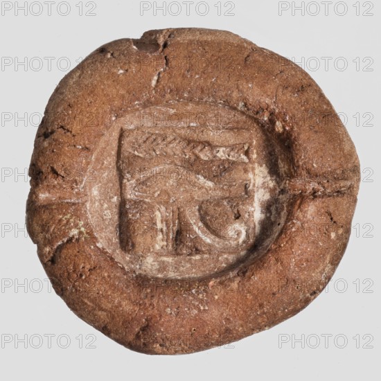 Mold for an amulet of the eye of Horus udjat. Hemispherical mold with a representation of the udjat impressed in the recess; two small channels lead from the recess to the edge. Castings; mold. Late Period, 664–332 BC, Egypt. Height 1.4 cm, width 3.5 cm, depth 3.5 cm., A round, flattened object with an uneven, textured surface in shades of brown and tan, featuring a shallow, oval depression in the center that contains carved or pressed linear and curved patterns with lighter beige highlights; the outer rim is irregular and slightly raised with small cracks and rough patches, and there are subtle variations of darker and lighter brown across the surface.