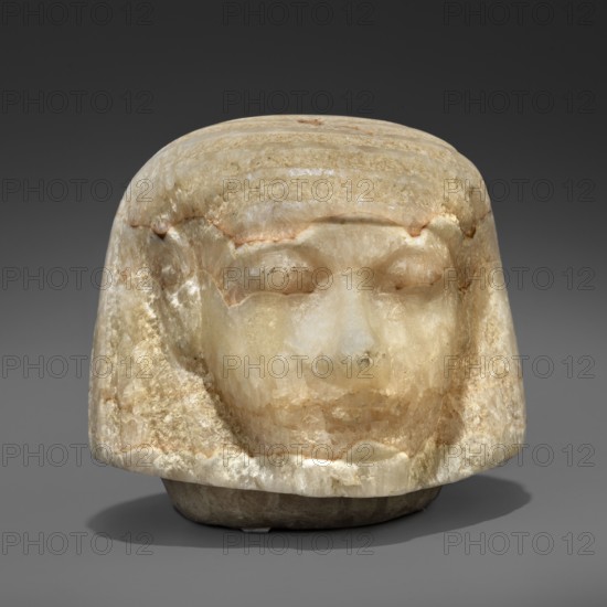 Canopic urn lid in the shape of a human head, Sons of Horus mythology, tomb equipment. This head is characterized by extremely subtle modelling, characteristic of the best works of the Amarna and early post-Amarna periods 2nd half of the 14th century BC. Delicate, aristocratic facial features, sfumato especially around the eyes, a wig with stripes of varied texture—features found in sculpture of the highest quality. c. 1350–c. 1295 BC; New Kingdom, end of 18th Dynasty, Egypt. Canopic urn. Height 8.2 cm; width 8.6 cm; depth 9.2 cm; weight 1.124 kg., A carved stone head with a rounded top and stylized hair, showing a smooth pale surface with a few areas of translucence and subtle brownish and beige veining; the facial features include closed eyes, a broad nose, and a faint mouth, all rendered in the same pale cream and off-white tones with warmer beige and light brown accents; the sides and lower edge show rougher, textured areas with creamy white and tan colors; the object sits on a dar...