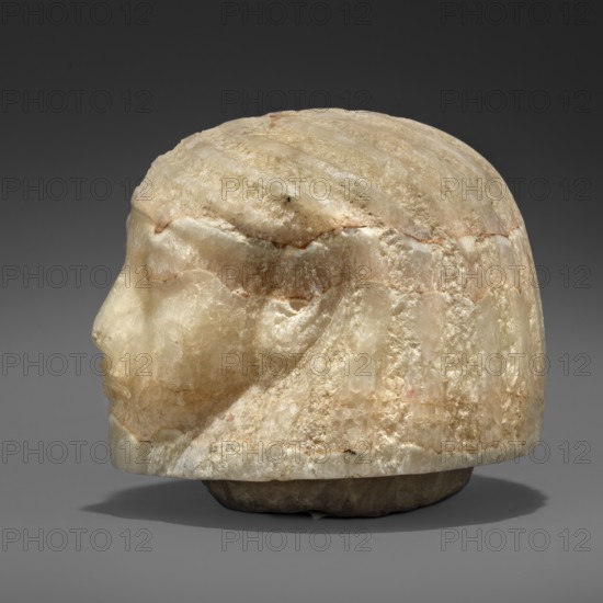 Canopic urn lid in the shape of a human head, Canopic urn lid, Sons of Horus mythology, tomb equipment. This head is characterized by extremely subtle modelling, characteristic of the best works of the Amarna and early post-Amarna periods 2nd half of the 14th century BC. Delicate, aristocratic facial features, sfumato especially around the eyes, a wig with stripes of varied texture—features found in sculpture of the highest quality. c. 1350–c. 1295 BC; New Kingdom, end of 18th Dynasty, Egypt. Height 8.2 cm; width 8.6 cm; depth 9.2 cm; weight 1.124 kg., A carved stone head shown in profile with a rounded top and stylized facial features, rendered in translucent pale tones including cream, beige, off-white, and hints of light brown and pink, with areas of rough crystalline texture and smoother polished surfaces; the hair or headdress is suggested by shallow incised lines across the top, and the piece rests on a darker base with gray and brown tones against a neutral dark gray background.
