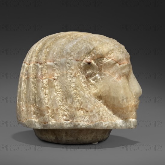 Canopic urn lid in the shape of a human head, Sons of Horus mythology; tomb equipment. This head is characterized by extremely subtle modelling, characteristic of the best works of the Amarna and early post-Amarna periods 2nd half of the 14th century BC. Delicate, aristocratic facial features, sfumato especially around the eyes, a wig with stripes of varied texture—features of high-quality sculpture of the period. c. 1350–c. 1295 BC; New Kingdom, end of 18th Dynasty, Egypt. Canopic urn. Height 8.2 cm; width 8.6 cm; depth 9.2 cm; weight 1.124 kg., A carved stone object resembling a head in profile sits against a dark gray background, showing a smooth rounded top and a stylized face with a prominent nose and chin; the surface of the stone displays a mix of creamy white, beige, pale yellow, and light brown tones with subtle translucent areas and banded striations, and there are a few rougher textured patches and thin reddish-brown lines running across the form, with a shadow visible benea...