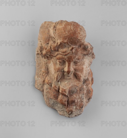 Mask of Bearded Dionysus. Miniature mask depicting Dionysus with an expressive nose, full lips in a slight smile framed by a mustache and a thick, curled beard. On the head with a lush mane of hair is a wreath, and above it a fragmentarily preserved and unidentified decoration. Fairly good level of workmanship. 2nd half of the 2nd century BC. Myrmekion place of discovery, Amissos place of origin? . Mask, height 7 cm., A fragment of a carved stone face with a textured, uneven surface, showing sculpted hair, eyebrows, eyes, a nose, and the suggestion of a beard or lower facial features; the stone displays a range of warm reddish-brown and terracotta tones with areas of lighter beige and some darker brown patches, set against a neutral light gray background.