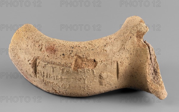 Fragment of a spout and vice from a Sinopean transport amphora with a three-line stamp depicting the head of Heracles and the Greek inscription When Demetrios was astynome made by Mithradates. The spout is a not very massive roller bent outward. The vice is oval in cross-section, slightly flattened. The first line of the stamp contains the astynomic titulary; the second names the astynomeus Demetrios I in the generic form; the third names the manufacturer Mithradates in the nominative. Head of Heracles facing left. Hellenistic period, 280–270 BC. Sinope origin. Stamp impressions; Sinopean stamp. Height 7.1 cm, width 5.3 cm, depth 9.3 cm., A curved, broken ceramic fragment rests against a smooth grey background; the fragment is a sandy beige color with darker brown and black speckles and a few reddish-brown smudges, showing a rough, porous texture and irregular edges where it has broken. The surface includes an indented rectangular area and shallow impressed marks, and one end of the fr...