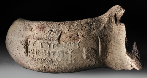 Fragment of a vice from a transport amphora with a stamp depicting the head of Heracles and the Greek inscription When Demetrios was astynome made by Mithradates., or workshop. Hellenistic period, Sinopean stamp on a stamped vice; head of Heracles iconographic; stamp impressions. Fragment of the spout and vice from a Sinopean amphora with the stamp of the astynome Demetrios I and the maker Mithradates. The spout is a not-very-massive roller bent outward. The vice is oval in cross-section, slightly flattened. Three-line stamp legend: first line contains the astynomic titulary; second line contains the name of the astynomeus Demetrios I in the generic form; third line contains the manufacturer Mithradates in the nominative. To the right of the legend is the head of Heracles, turned left. Attributed to M. Matera; dated 280–270 BC. Findspot: Myrmekion; origin: Sinope. Stamp, vice; height 7.1 cm, width 5.3 cm, depth 9.3 cm., A weathered curved ceramic fragment rests against a dark backgroun...