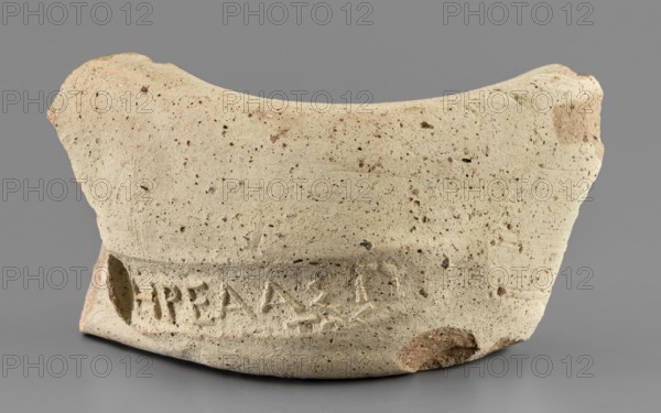 Fragment of a vice behind a stamp, 300–275 BC, Chersonesos Chersonesos Taurica; found at Myrmekion. Stamp; height 3.3 cm, width 5.5 cm, length 5.5 cm., A fragment of a pottery rim or vessel displayed against a neutral gray background, made of a beige to light tan coarse clay with numerous small dark brown and reddish inclusions and pitting across its surface, showing an irregular curved top edge and a thicker lower band with a shallow recessed area, a few rougher brownish patches where the surface is chipped, and subtle horizontal tool or wheel marks visible on the lower portion.