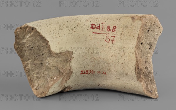 Fragment of a vice behind a stamp, material from excavations, 300–275 BC, Chersonesos Chersonesos Taurica place of origin, Myrmekion place of finding, stamp, height 3.3 cm, width 5.5 cm, length 5.5 cm, A broken curved ceramic fragment with a smooth pale beige surface and rougher brown edges, showing a few small speckles and granular textures; a group of reddish marks appears on the pale area; the background is a flat medium gray.