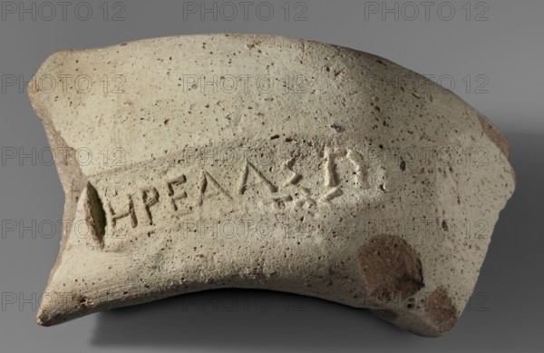 Fragment of a vice behind a stamp, material from excavations, 300–275 BC, Chersonesos Chersonesos Taurica place of origin, Myrmekion place of finding, stamp, height 3.3 cm, width 5.5 cm, length 5.5 cm, A curved fragment of pottery with a rough, pitted surface in shades of beige, light tan, and off-white, showing several small darker brown speckles and a few larger brown patches; the piece has irregular broken edges with exposed inner material that is darker brown, and shallow incised marks across its face; the background is a smooth gray.