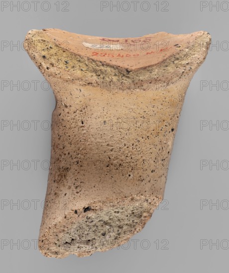 Fragment of a vice from a Sinopean transport amphora with a rectangular four-line stamp: astynomenic title on the first line; astynomeus' name and patronymic Posis, son of Stratonikos on the second and third lines; the fourth line name of the amphora's manufacturer not impressed. To the right of the legend, at the height of the second and third lines, an emblem in the form of a helmeted head of Athena. Inscription reading Made when Posios son of Stratonicus was astynomeus. Dating: 320–300 BC previously dated ca. 150–100 BC. Excavated at Myrmekion Crimea; Sinopean origin. Stamp; fragment dimensions: height 7.2 cm, width 6 cm, depth 2.3 cm., A fragment of a ceramic object with a rough, porous surface visible against a neutral gray background, showing a warm beige to light tan body with darker speckles and small inclusions throughout, a rim area with a slightly pinkish-orange inner surface and a rough, crumbling edge that has patches of pale yellow and off-white, and a broken end revealin...