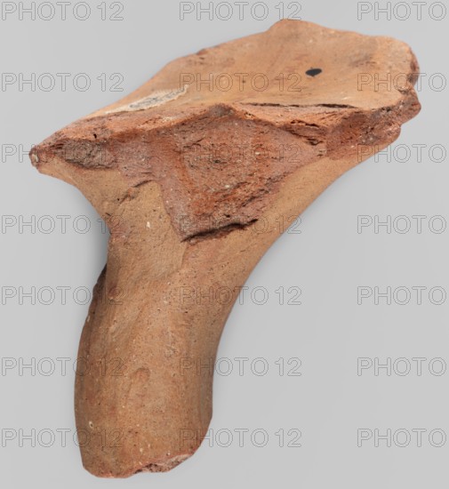 Fragment of a stamped amphora neck, 332–30 BC; Hellenistic period, Knidos Caria, Myrmekion place of finding, amphora neck, stamp; height 6.5 cm, width 7.7 cm, depth 6.5 cm, length 6.5 cm, A fragment of a hard, irregular object with a curved protruding section and a flatter, broken top surface, shown against a plain light gray background. The object displays a range of earthy tones including reddish-brown, terracotta, and sandy beige, with areas of darker brown and small pale speckles. The surfaces vary in texture from rough, crumbly-looking broken edges to smoother, slightly pitted curved areas. There are a few small chips and indentations along the edges and one small dark mark on the flatter top surface.