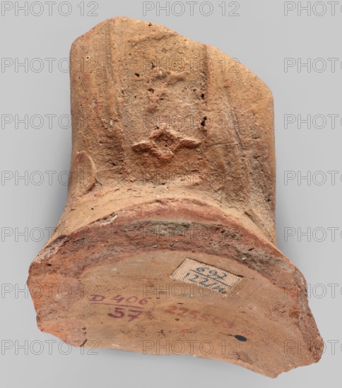 Fragment of a stamped amphora neck, 332–30 BC; Hellenistic period, possibly Knidos Caria, found in Myrmekion; amphora neck with stamp; height 6.5 cm, width 7.7 cm, depth 6.5 cm, length 6.5 cm., A broken terracotta fragment with a rough, weathered surface in shades of orange, reddish-brown, and tan rests against a light gray background; the fragment shows a curved rim and a flat base area with worn edges, pitted texture, and a raised decorative motif on one side, and there is a small rectangular patch of a lighter beige fabric-like material adhered to the base.