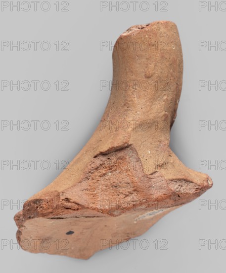 Fragment of a stamped amphora handle, 332–30 BC; Hellenistic period, Knidos Caria, Myrmekion finding. Handle, stamp; height 6.5 cm, width 7.7 cm, depth 6.5 cm, length 6.5 cm, A fragment of a broken object with a rough, uneven surface in shades of tan and reddish-brown, showing textured and smoother areas, with some darker brown and pale beige speckles and a small dark spot; the piece has an irregular, angular shape with a thicker rounded section and a thinner flared edge, visible chips and pitted areas, and subtle shadowing against a light gray background.