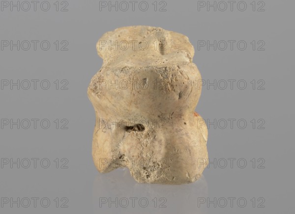 Cow astragal, Myrmekion Crimea, Hellenistic period, antiquity, cow astragal condylo-talus bone, 332–30 BC, Myrmekion place of finding, product of nature, height 3.4 cm, A single irregularly shaped object with a rough, pitted surface sits centered against a flat background; the object is primarily beige with areas of darker tan and pale ochre, and it shows textured grooves, small cavities, and uneven edges. The background is a uniform muted gray. There is a subtle reflection of the object on the smooth surface beneath it. The object’s surface displays speckled darker spots and slightly smoother worn areas, and one side shows a small concave indentation.