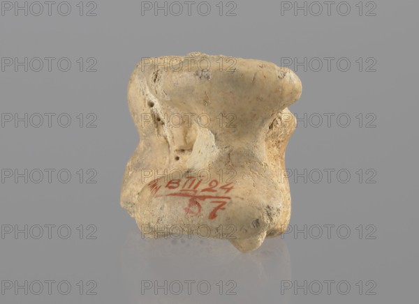Cow astragal, Myrmekion Crimea, Crimea Ukraine, Hellenistic period, antiquity, bones, cow astragal condylo-talus bone, 332–30 BC, Myrmekion place of finding, product of nature, height 3.4 cm, A small irregularly shaped object with a rough, porous surface, shown against a uniform gray background and resting on a reflective surface. The object is primarily beige and light tan with areas of darker tan and light brown discoloration and small pits and indentations. It has rounded protrusions and concave depressions, with a few smoother worn areas and some rougher, crumbly textures. There is a visible mark in a red color on one side of the object. The reflection of the object on the surface below shows muted beige and gray tones.