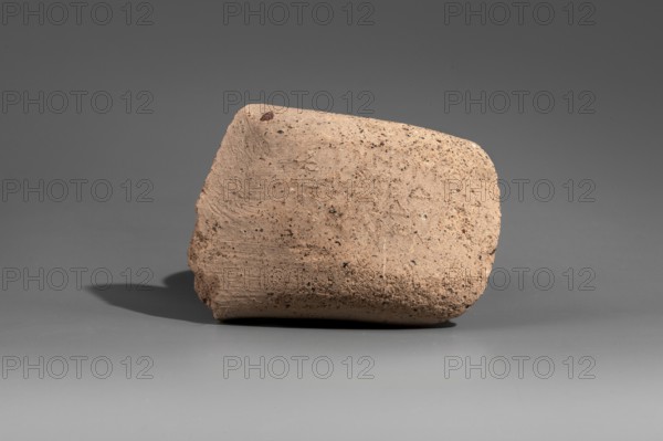 Fragment of a stamped amphora vice, Cephalion ca. 260–205 BC. Fragment of a Sinopean amphora vice with the stamp of the astynomeus Leomedon son of Epidemos and the manufacturer Cephalion. A four-line rectangular stamp on the upper surface: first line the astynomenic title; second and third lines the astynomeus' name and patronymic; fourth line the manufacturer's name in the genitive. 2nd half of the 3rd century BC. Sinope origin; Myrmekion Crimea findspot. Stamp; vice; height 6.8 cm., A roughly rectangular piece of pottery or stone rests on a smooth surface, showing a textured, grainy surface with irregular edges and small pits; the object is primarily beige with speckles of darker brown and occasional tiny black flecks, and it casts a soft shadow on the gray background and tabletop, which are shades of medium and light gray respectively.