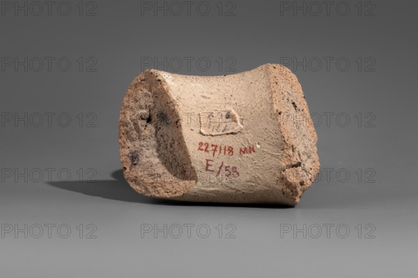 Fragment of a stamped amphora vice, Cephalion ca. 260–205 BC. Fragment of a Sinopean amphora vice with the stamp of the astynomeus Leomedon son of Epidemos and the manufacturer Cephalion. A four-line rectangular stamp on the upper surface: first line the astynomenic title; second and third lines the astynomeus' name and patronymic; fourth line the manufacturer's name in genitive. 2nd half of the 3rd century BC; Sinope origin; Myrmekion Crimea finding. Stamp, vice; height 6.8 cm., A roughly cylindrical, broken clay object sits on a flat surface, showing a textured, somewhat porous exterior with irregular edges and chipped areas; the clay appears in shades of beige, tan, and light brown with darker brown speckles, and there is a small patch of frayed fabric stuck to one side in pale off-white and beige tones; the background and surface are smooth and gray, and a soft shadow in darker gray lies beside the object.