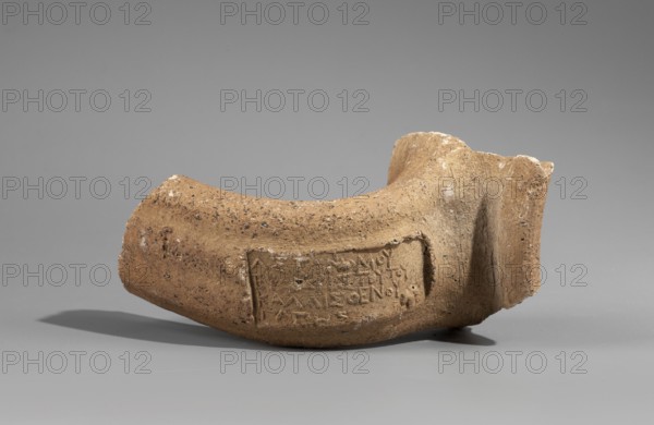 Fragment of the spout and vice of an amphora with a stamp Papes, c. 255–205 BC. Hellenistic Sinopean amphora: preserved fragment of the spout, neck, and stamped vice. Spout formed as a semicircular roller; vice oval in cross-section, flattened. Rectangular four-line stamp; to the right of the legend at level of lines 3–4 an emblem in the form of a grapevine. First line: astynomic titulary. Second line: name of the astynomeus Eucharistos in generic form. Third line: his patronymic. Fourth line: name of the manufacturer Papes in generic form. 2nd half of the 3rd century BC. Findspot: Myrmekion Crimea. Height 9.2 cm., A broken curved ceramic fragment rests on a smooth gray surface against a gray background; the fragment is an earthy brown color with darker brown speckles and rough textured areas, showing a recessed rectangular panel on one side and an uneven broken edge on the other, with a shadow cast beneath it.