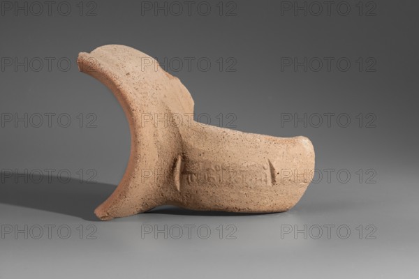 Fragment of the spout and stamped vise of a Sinopean amphora. The spout is a low, semicircular cylinder; the vise is oval in cross-section. On its upper surface is a partially impressed stamp of the astynomas Femios, son of Theopeithos. The first line of the legend the astynomas' titulary is missing; the second and third lines contain the astynomas' name and patronymic. To the right of the legend is an emblem showing a man trampling grapes placed in a basket. 2nd half of the 3rd century BC. Sinope origin. Height 7.9 cm; width 10.7 cm., A fragment of a textured terracotta object rests on a smooth surface, showing a curved rim and a thicker cylindrical section with worn edges; the object is a light brown to beige color with small darker speckles, and it casts a soft shadow on the surface; the background and surface are shades of gray and darker gray.