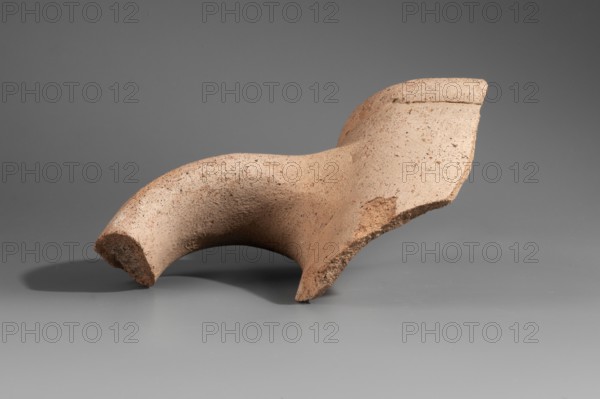 Fragment of the spout and stamped vise of a Sinopean amphora. The spout is a low, semicircular cylinder; the vise is oval in cross-section. On its upper surface is a partially impressed stamp of the astynomas Femios, son of Theopeithos. The first line of the legend the astynomas' titulary is missing; the second and third lines contain the name and patronymic. To the right of the legend is an emblem showing a man trampling grapes placed in a basket. 2nd half of the 3rd century BC. Sinope. Height 7.9 cm; width 10.7 cm., A broken curved fragment of a clay or ceramic object rests on a smooth surface; the fragment is a light tan or beige color with darker brown speckles and rough, grainy texture on some edges, and it casts a soft gray shadow on the flat gray background and surface.