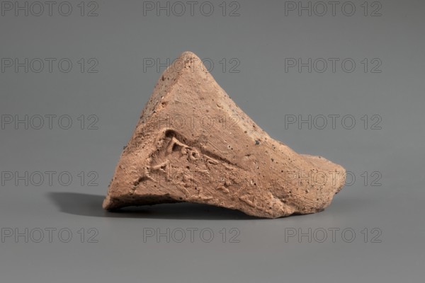 Fragment of a Sinopean amphora vice with the stamp of the astynomeus Poseidonios son of Aeschinos and the manufacturer Kallisthenes ca. 290–280 BC. Only fragments of the first and second lines of the legend have survived: the first line contained the astynomeus’s name; the second contained the patronymic and the title in abbreviated form; the third originally contained the manufacturer’s name, Kallisthenes. Sinopean amphora fragment, stamp; height 5.7 cm; findspot: Myrmekion Crimea., A triangular fragment of reddish-brown clay with a rough, pitted surface sits on a smooth gray background. The clay piece shows textured tool marks and indentations, with areas of darker speckling and small holes. Shadows fall beneath and to the left of the fragment, and the overall color palette includes reddish-brown, gray, darker brown specks, and muted shadow tones.