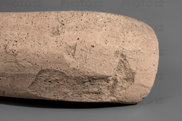 Fragment of a Sinopean amphora vice with a partially preserved rectangular stamp. The stamp is oval in cross-section, flattened, and located on the bend of the vice. Originally a four-line stamp: the first line of the legend is not impressed; the last line is partially preserved only upper parts of letters visible; last two letters missing. To the right of the legend was an emblem in the form of a prora. Originally, the first line contained the astynomic title; the second line contained the name of the astynomeus Delphios in the generic form; the third line is his patronymic; the fourth line contains the name of the manufacturer Agathon II in the nominative. Maker: Agathon II ca. 250–205 BC. Date: 2nd half of the 3rd century BC. Findspot: Myrmekion Crimea. Origin: Sinope. Type: stamped amphora vice stamp, vice. Height: 12.8 cm., A close-up of a cylindrical object with a rough, sandy surface in shades of tan and light brown dotted with small dark specks, resting on a smooth grey backgro...