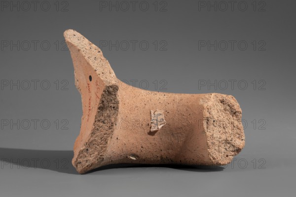Fragment of a Sinopean amphora vice with a rectangular stamped impression inscription reversed, read right to left preserving the name of the Sinopean manufacturer Sagaris with patronymic; an emblem in the form of a leaf originally stood at the left of the stamp. Hellenistic period, ca. 320–300 BC. Found at Myrmekion Crimea. Stamp; height 8 cm. Made in Sinope. Maker attributed: Sagaris son of Numenius., A broken terracotta fragment rests on a flat surface against a neutral gray background, showing rough, porous edges and smoother inner surfaces. The piece is primarily a warm orange-brown color with darker speckles scattered across its surface and a few areas of lighter, beige-toned roughness where the ceramic is fractured. A small patch of fabric-like material is attached to the smoother face, displaying a mix of off-white and darker threads. Shadows beneath the fragment are muted gray, and there is a small round dark spot visible on one side of the piece.