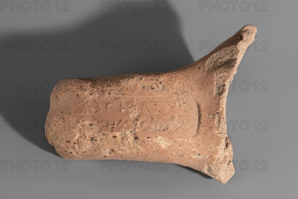 Fragment of a Sinopean amphora vice with a rectangular stamp impression on the upper surface. The fragmentarily preserved, reversed inscription read right to left contains the name of the Sinopean manufacturer, Sagaris, given with his patronymic; an emblem in the form of a leaf originally appeared at the left of the stamp. Dated ca. 320–300 BC. Origin: Sinope; findspot: Myrmekion. Height 8 cm. Maker: Sagaris son of Numenius., A fragment of a rough, earthen object with a textured, pitted surface in shades of tan and light brown rests against a smooth gray background; the object has an irregular, slightly cylindrical form with a flared, broken end and a rounded opposite end, showing a mix of darker speckles and small porous holes across its surface and casting a soft shadow on the background.