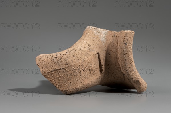 Fragment of a stamped amphora neck, Kinolis c. 290–250 BC. Fragment of a Sinopean amphora neck with the stamp of the astynomeus Attalos and the manufacturer Kinolis. Rectangular stamp with a three-line legend: first line the astynomeus's name in genitive; second line the astynomean title; third line the manufacturer's name in genitive. To the right of the legend, an emblem of a male head in profile facing right. Male heads iconography. 284–280 BC. Myrmekion findspot. Sinope origin. Stamp; neck; height 7.5 cm., A fragment of a rough-textured pottery piece rests on a smooth surface against a plain background; the pottery is a warm tan color with darker speckled spots and areas of lighter discoloration, showing an uneven, grainy surface and chipped edges, with a curved rim and a hollow interior visible at one end, casting a soft shadow beneath it.