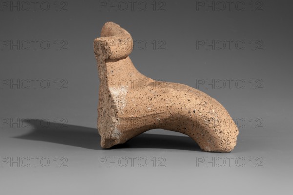 Fragment of a stamped amphora neck, Kinolis c. 290–250 BC. Fragment of a Sinopean amphora neck with the stamp of the astynomeus Attalos and the manufacturer Kinolis. Rectangular stamp with a three-line legend: first line contains the astynomeus's name in the genitive; second line the astynomean title; third line the manufacturer's name in the genitive. To the right of the legend, an emblem in the form of a male head in profile, turned to the right. Attributed to M. Matera, dated 284–280 BC. Findspot: Myrmekion; origin: Sinope. Stamp; neck; height 7.5 cm., A small sculptural object with a rough, speckled tan surface sits on a smooth gray surface against a darker gray background; the object has a curved, arching form with a thicker upright section and a rounded protrusion, showing irregular texture with darker flecks and patches of lighter wear, and it casts a soft shadow to the left.