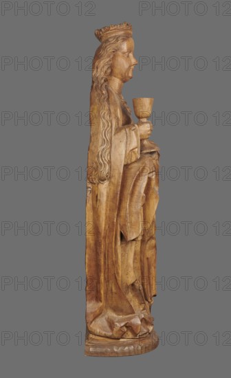 Figure of St. Barbara, medieval wall sculpture, hollowed out from behind head also has a circular hollow. Depicts St. Barbara in contrapposto, creating a slight S-shaped silhouette. Slightly oversized head on a cylindrical neck; elongated face with high forehead, straight narrow nose, full cheeks, small smiling mouth with dimples and a dimpled chin. Long hair framed and falling down her back, one strand draped over her right shoulder, carved in thin parallel wavy strands. She wears a crown finial broken. Right hand grasps the stem of a chalice; left hand, fingers covered by a cloak, supports the foot of the chalice. Dressed in a gown with triangular neckline and long wide sleeves, cinched with a decorative belt; voluminous cloak with numerous folds and dry thin ridges. Pointed left shoe peeks from beneath the folds. Round, irregularly carved base. No polychrome. ca. 1500, possibly Silesia. Sculpture, height 102.0 cm, width 35.0 cm, depth 24.0 cm., A wooden carved figure shown in profil...