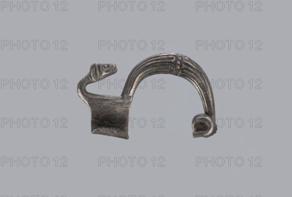 Snake-headed fibula, Southern Italian workshop, fibulae, Italy, snake iconography, silver. Fibula with an arched, thick hoop. The hoop is terminated on one side with an imitation spiral, and on the other side connected to the rectangular wall of the sheath, bent at the bottom. A cylindrical leg extends from the sheath, bent towards the hoop, ending with a snake's head. The hoop is decorated with numerous longitudinally engraved lines. Condition: corrosion, missing needle. 5th–4th century BC, Southern Italy., A small metallic object resembling a curved clasp or decorative piece sits against a plain gray background, showing a dark silver color with areas of lighter silver highlights; one end features a stylized animal head with a slightly open mouth and ridged neck, the middle has a flat rectangular section with a textured, worn surface, and the opposite side forms an arched, ribbed band that terminates in a rounded, curled end.