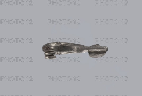 Snake-headed fibula, Southern Italian workshop. Fibula with an arched, thick hoop terminated on one side with an imitation spiral and on the other connected to the rectangular wall of the sheath, bent at the bottom. A cylindrical leg extends from the sheath, bent towards the hoop, ending with a snake's head. The hoop is decorated with numerous longitudinally engraved lines. Material: silver. Condition: corrosion, missing needle. Date: 5th–4th century BC. Iconography: snake. Type: fibula., A small metallic object with a dull silver-gray surface rests against a plain light gray background; the object has an elongated central body with a slightly rounded end and a flattened end, a few raised ridges and grooves along its length, and a small curl or hooked projection at one side, showing areas of smoother and rougher texture and subtle darker gray tarnish.