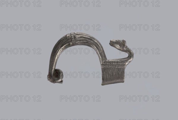 Snake-headed fibula, unknown Southern Italian workshop. Fibula with an arched, thick hoop. The hoop is terminated on one side with an imitation spiral, and on the other side connected to the rectangular wall of the sheath, bent at the bottom. A cylindrical leg extends from the sheath, bent towards the hoop, ending with a snake's head. The hoop is decorated with numerous longitudinally engraved lines. Silver. Condition: corrosion, missing needle. 5th–4th century BC, Southern Italy. Snake iconography., A metallic curved object with a patinated silver-gray surface sits against a plain light gray background; the object has a smooth arching band with ridged grooves along its outer curve ending in a small curled tip, and the opposite end forms a flattened rectangular section decorated with incised triangular patterns and a raised textured band, while a sculpted animal-like head with a tapered snout and folded features projects from one side, showing variations of dark gray, silvery highlight...