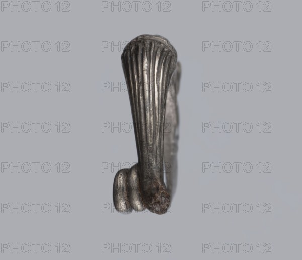 Snake-headed fibula, unknown Southern Italian workshop. Fibula with an arched, thick hoop. The hoop is terminated on one side with an imitation spiral, and on the other side connected to the rectangular wall of the sheath, bent at the bottom. A cylindrical leg extends from the sheath, bent towards the hoop, ending with a snake's head. The hoop is decorated with numerous longitudinally engraved lines. Condition: corrosion, missing needle. 5th–4th century BC, Southern Italy. Materials: silver. Subject/iconography: snake., A close-up of a small metallic object with a vertical ribbed surface and a rounded end, shown against a plain light gray background; the metal appears silver-gray with darker gray areas and a small patch of brownish rust or corrosion near one end, and there are a few curved ridges at the lower end of the piece.