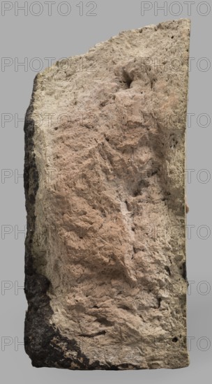Fragment of a brick with relief decoration. Element of an unglazed panel with a depiction of a bull from the decoration of the Ishtar Gate. On the face side, plastic decoration with a depiction of two diagonally running spirally twisted curls of hair from the figures of bulls that decorated the walls of the Ishtar Gate in Babylon older unglazed phase. 1st half of the 6th century BC; Neo-Babylonian period. Babylon Iraq. Element of architectural decoration. Weight 810 g., A rough, vertically oriented slab of stone with an irregular, textured surface and visible pits and crevices, showing areas of pale beige, pinkish-beige, light tan, dark brown, and gray; the left edge has darker brown to black patches while the central surface has more pinkish-beige tones and the right edge appears lighter beige, all set against a plain gray background.