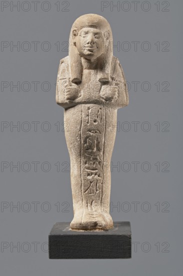 Ushabti, Mummy-shaped figurine made of limestone. Arms crossed on the chest, right folded over left, holding hoes in each hand. Rectangular basket on the back, slung by a single string over the left shoulder. Three-part smooth wig, almond-shaped eyes, wide nose, prominent mouth in a slight smile. Engraved inscription in a single column on the front of the legs; name illegible. Inscription reads: Shd Osiris mistress of the house of Naa, justified by her voice. Ca. 1295–1186 BC; New Kingdom, 19th Dynasty, Egypt. Shabti; height 13.3 cm, width 4.3 cm., A carved standing figure wearing a long wig with side locks, arms crossed at the chest holding small objects, and a long skirt that reaches the feet, displayed on a rectangular base; the figure is made of a light beige stone with surface texture and weathering, and the base is dark gray to black; the background is a uniform medium gray.