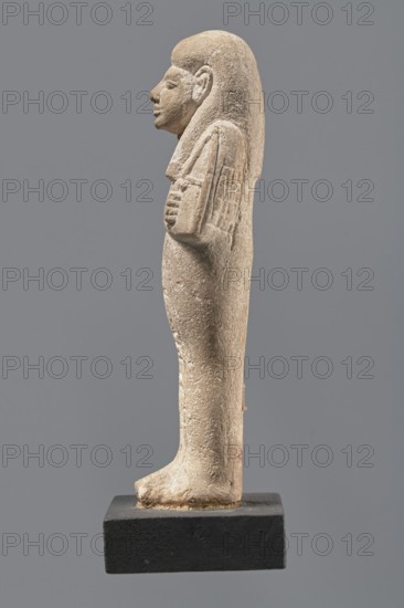 Ushabti, Mummy-shaped figurine made of limestone. Arms crossed on the chest, the right over the left, holding hoes in each hand. Rectangular basket on the back, slung by a single string over the left shoulder. Three-part smooth wig; almond-shaped eyes, wide nose, prominent mouth with a slight smile. Engraved hieroglyphic inscription in a single column on the front of the legs; name illegible. Inscription: Shd Osiris mistress of the house of Naa, justified by her voice. ca. 1295–ca. 1186 BC; New Kingdom, 19th Dynasty, Egypt. Shabti, height 13.3 cm, width 4.3 cm., A beige stone statuette of a standing human figure shown in profile, with a smooth rounded wig or hair extending down the back, a defined nose and chin, and a carved ear, arms folded across the chest with hands visible, textured carved details on the upper arms and torso, and a tapered lower body and legs ending in feet that rest on a rectangular dark base; visible colors include beige, light brown, and dark gray.