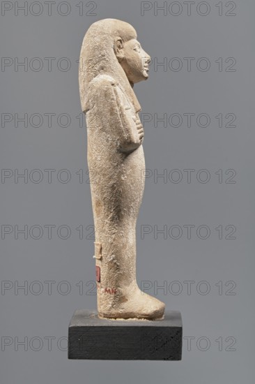 Ushabti, Mummy-shaped figurine made of limestone. Arms crossed on the chest, the right folded over the left, holding hoes in each hand. Rectangular basket on the back, slung by a single string over the left shoulder. Three-part smooth wig; almond-shaped eyes, wide nose, prominent mouth with a slight smile. Engraved inscription in a single column on the front of the legs; name illegible. Inscription text: Shd Osiris mistress of the house of Naa, justified by her voice. ca. 1295–ca. 1186 BC; New Kingdom, 19th Dynasty, Egypt. Shabti, height 13.3 cm, width 4.3 cm., A weathered stone figurine of a standing human figure shown in profile on a dark rectangular base, with a rounded head and a wig or hair that falls to the shoulders, a pronounced nose and chin, arms crossed over the chest, and simplified legs and feet. The stone surface appears rough and pitted with a worn texture. Visible colors include beige and light tan on the figurine, dark gray on the background, and black on the base, wit...