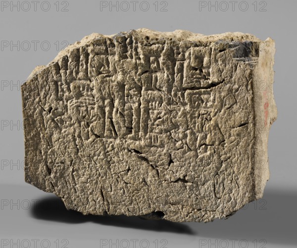 Fragment of a burnt clay brick with fragments of 7 lines of a cuneiform inscription, impressed with a stamp, of the Neo-Babylonian king Nebuchadnezzar II reign 604–562 BC. Archaized graphic style of cuneiform characters. Associated with Esagila Babylon and Ezida Babylon; origin likely Babylon or Borsippa, Southern Mesopotamia Iraq. Brick; weight 940 g; dimensions 15.1 cm × 12.4 cm × 4.9 cm., A roughly rectangular, irregularly shaped stone fragment sits against a plain gray background, showing a textured, weathered surface with many shallow grooves, cracks, and raised areas. The stone displays an overall beige to light tan color with areas of darker brown in crevices and slight grayish tones near the edges. The top edge is uneven and chipped, and one vertical side shows a smoother, lighter face with faint discoloration. The surface patterning forms a network of fissures and reliefs that create a rough, pitted appearance, and a soft shadow appears beneath the stone.
